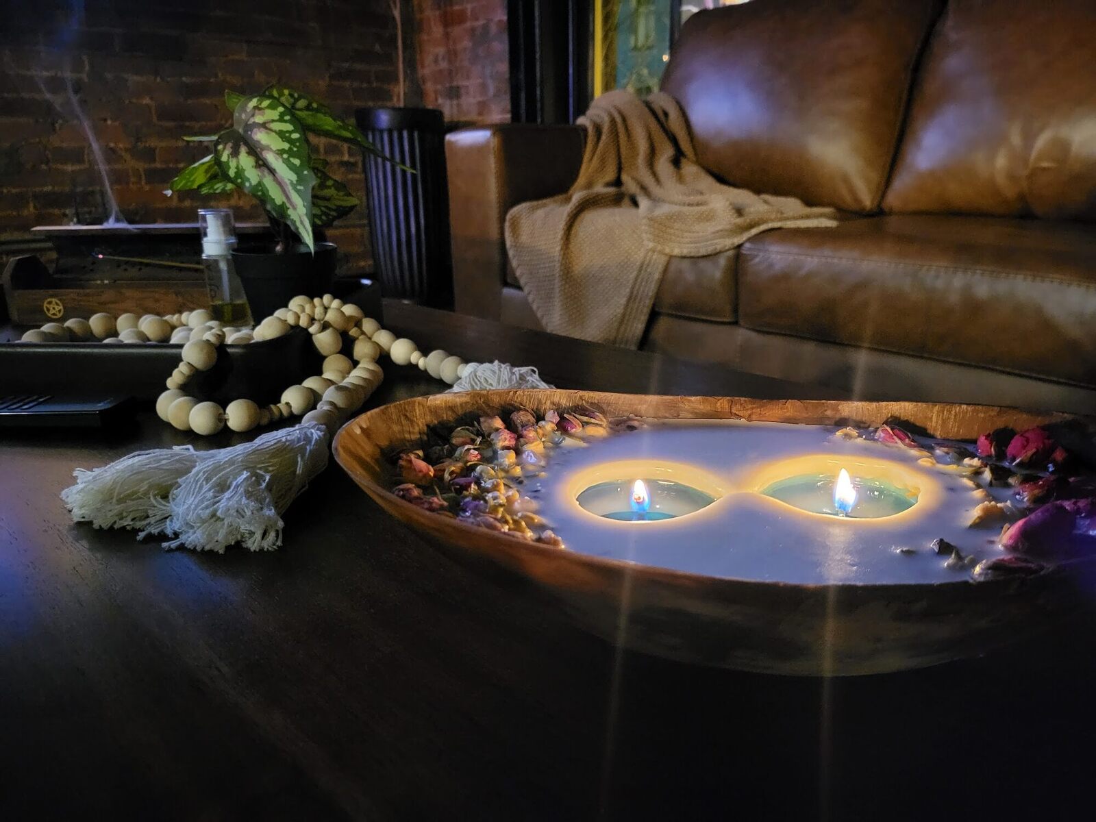 Two candles burning in a carved wooden bowl, dried petals around them, a leather sofa behind.