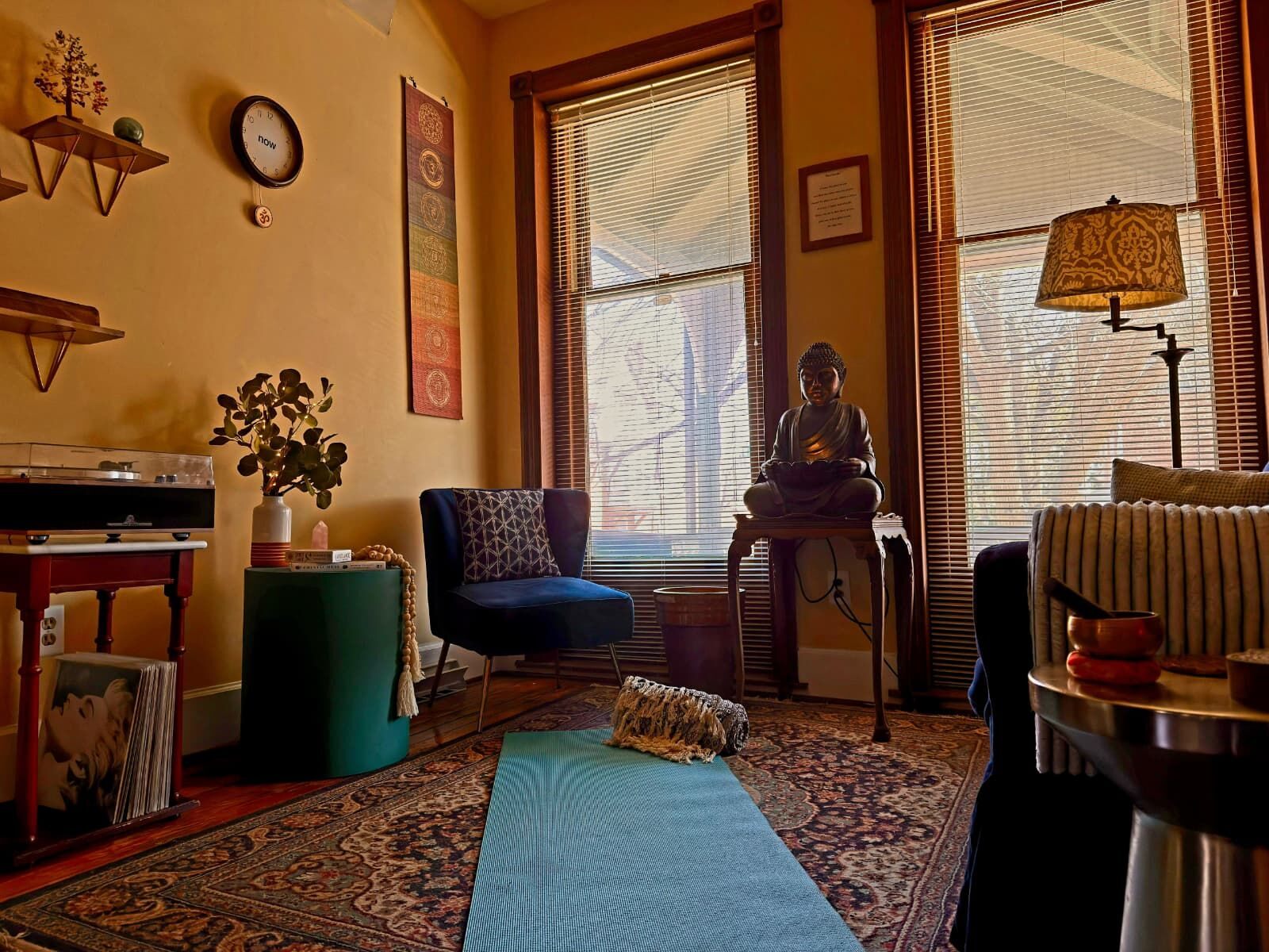 A yoga mat rolled out on an antique rug in morning light, a blue velvet chair and a Buddha by the tall windows.