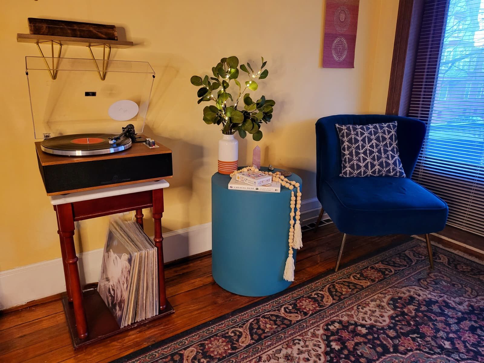 A record player on a wooden stand, records beneath it, a blue chair by the window.