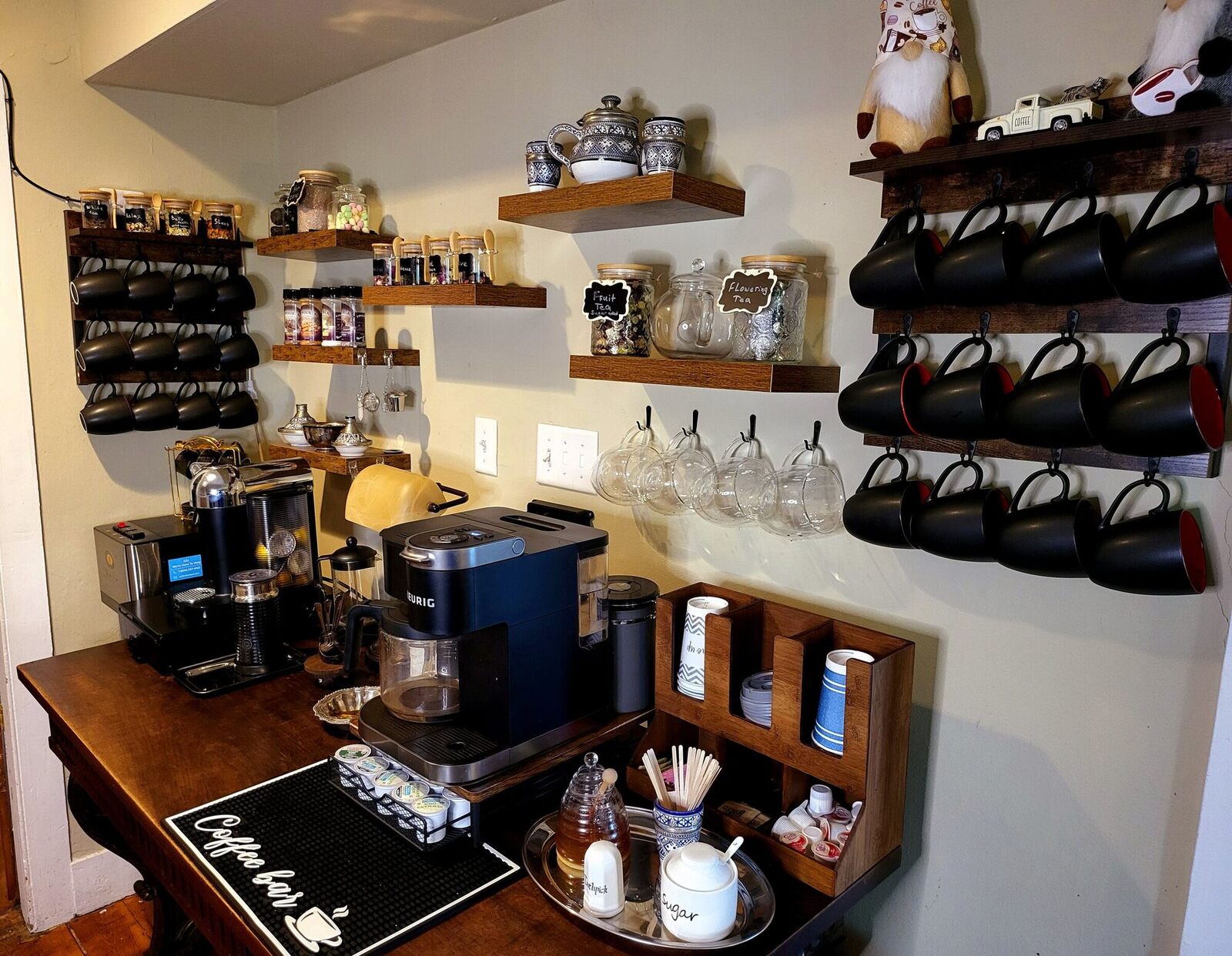 An antique table set with coffee and tea, black cups on a wall rack above it.