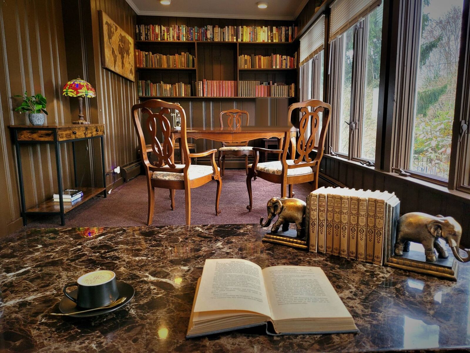A small library lined with old books, carved wooden chairs round a table by the windows, a stained-glass lamp lit in the corner, and a cup of coffee beside an open book.