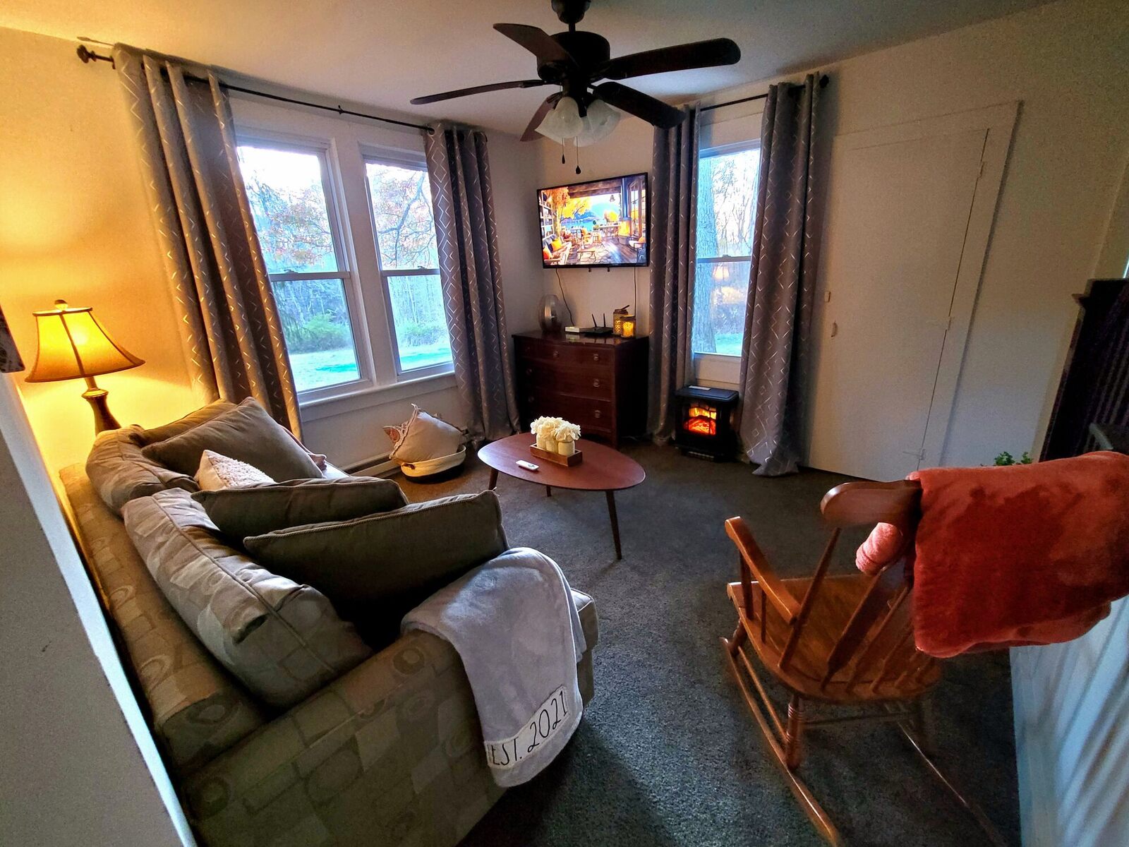 The family cottage's sitting room at dusk, a lamp lit beside the sofa, a rocking chair with a red throw and a small stove glowing by the window.