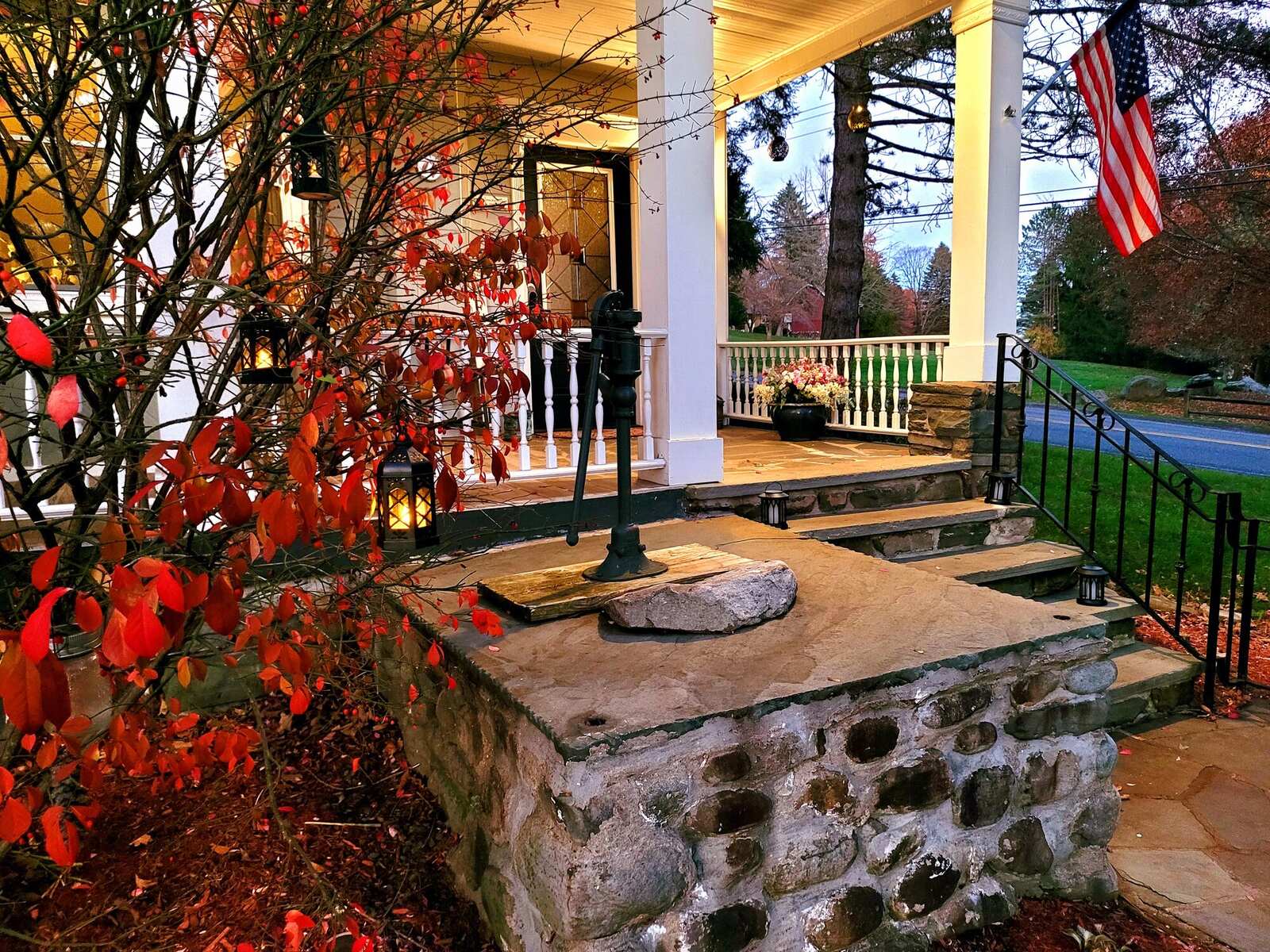 The original well in front of the Pump House, an iron hand pump on its fieldstone top, lanterns lit along the porch at dusk.