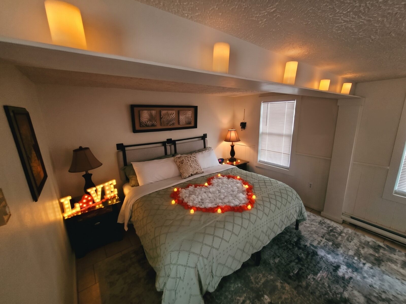 A suite set for a couple: a heart of rose petals ringed with small candles on the bed, a lit LOVE sign on the nightstand and candles along a high shelf.