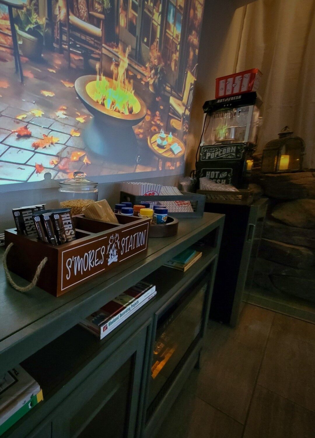 A film glowing on the wall above a s'mores station, jars of popcorn kernels and a popcorn machine beside a lantern.
