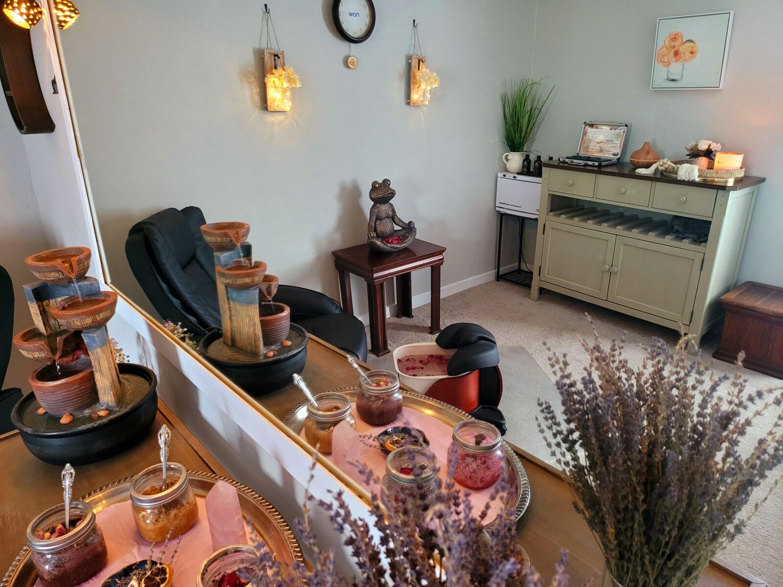 The Pump House spa room: a foot bath scattered with rose petals before a reclining chair, soak jars and a rose-quartz point on a silver tray, dried lavender, and on the wall a clock whose face reads NOW.