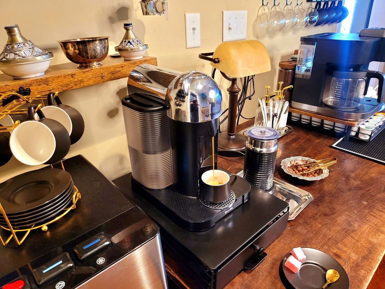 Coffee pouring at the Tea & Coffee Atelier in the Pump House foyer, cups stacked beside the machine and a lamp lit on the antique table.