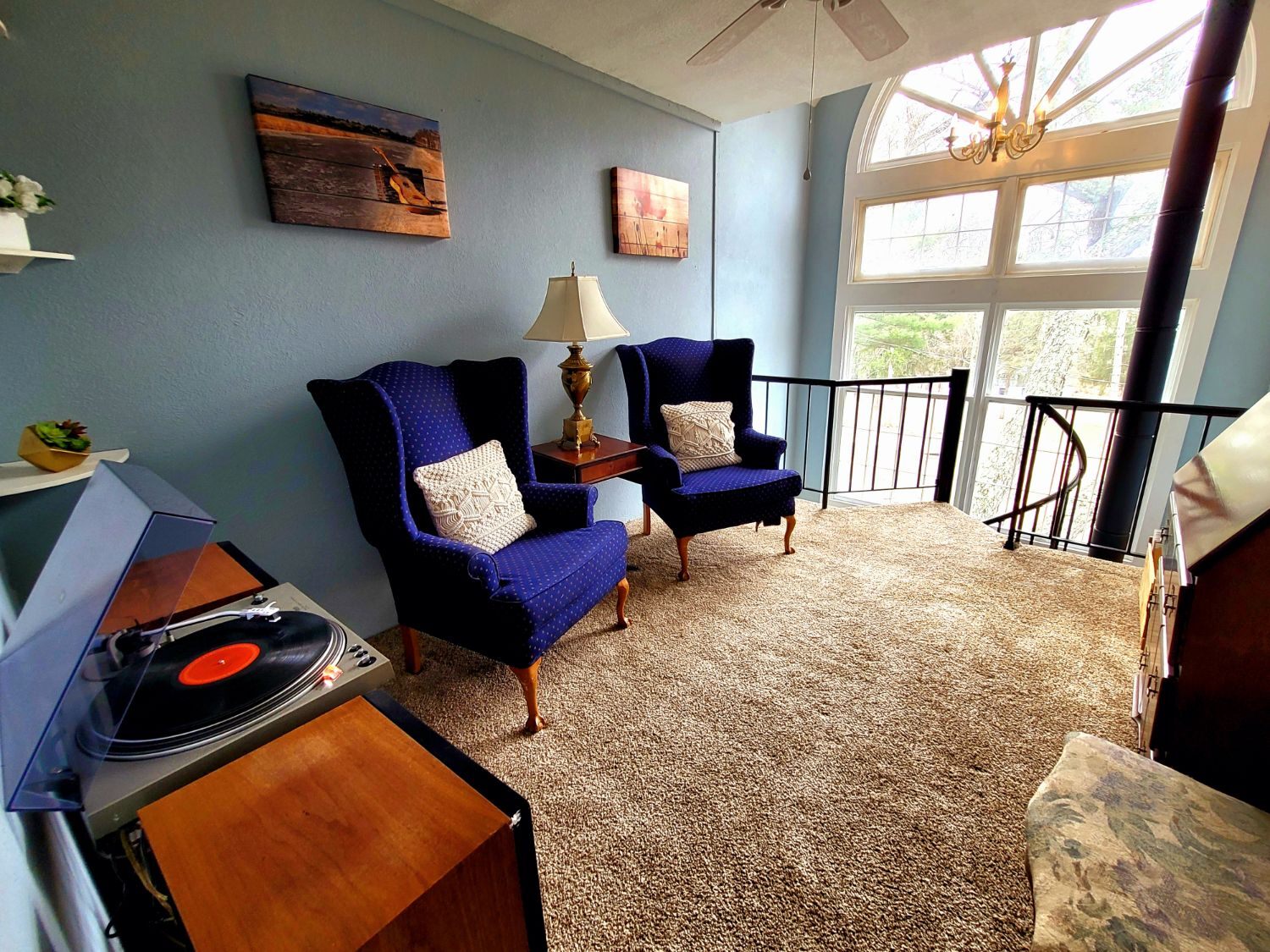The loft at the Pump House: a record playing on a turntable, two blue wingback chairs beside a brass lamp, and a tall arched window over the stair.
