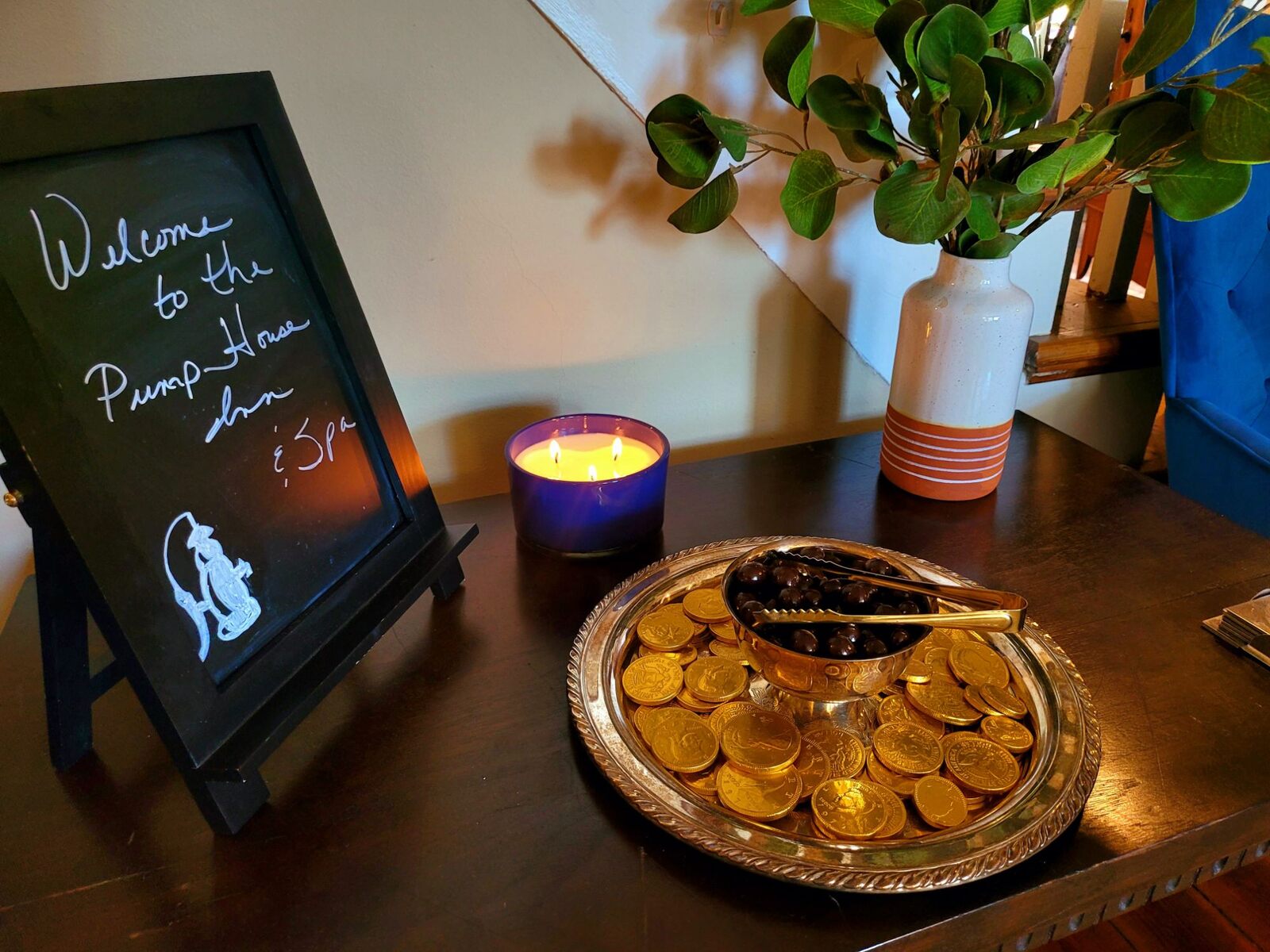 A three-wick candle lit on the foyer table beside a chalkboard that reads Welcome to the Pump House Inn & Spa, and a silver tray of chocolate coins and espresso beans.